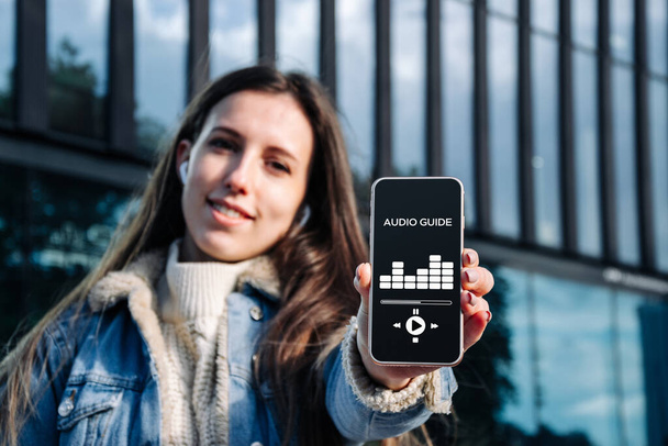 Audio tour online app on digital mobile smartphone. Happy young student woman holding phone listening audioguide. Simultaneous translation devices - Fotó, kép