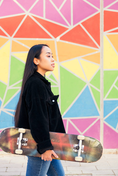 young asian woman walking with a skateboard in her hand in front of a colorful wall - Φωτογραφία, εικόνα