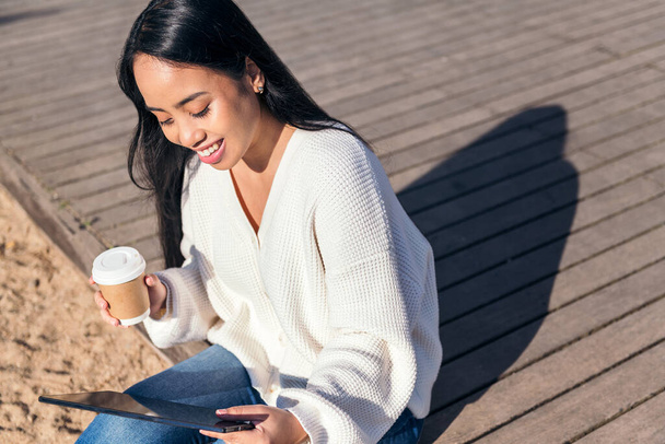 młoda Azjatka uśmiecha się siedząc na zewnątrz i patrząc na tablet z kawą na wynos w ręku - Zdjęcie, obraz