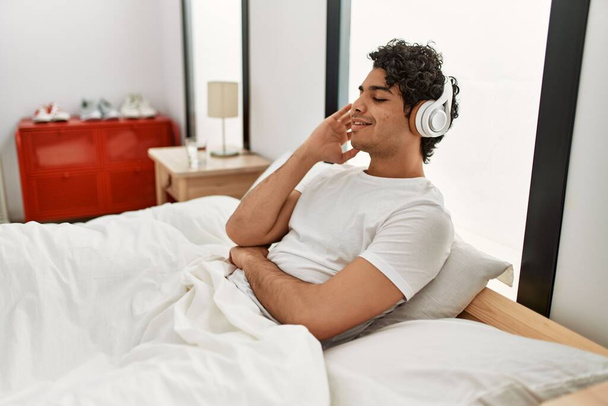 Young hispanic man listening to music sitting on the bed at bedroom. - Fotó, kép