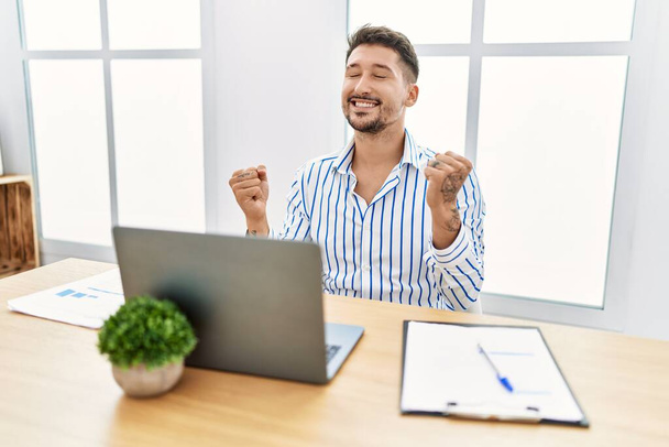 Young handsome man with beard working at the office using computer laptop very happy and excited doing winner gesture with arms raised, smiling and screaming for success. celebration concept.  - Photo, image
