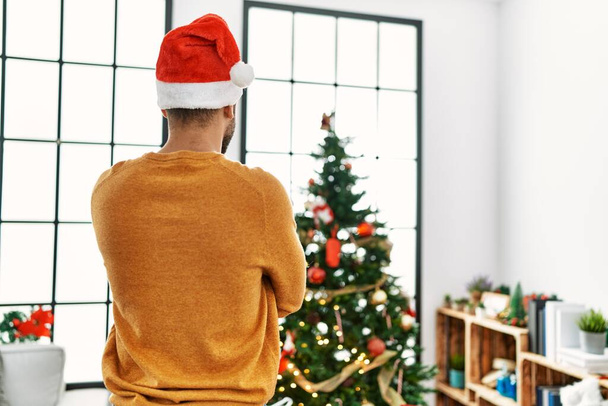 Hombre árabe joven en la vista trasera de pie con los brazos cruzados gesto por el árbol de Navidad en casa. - Foto, imagen