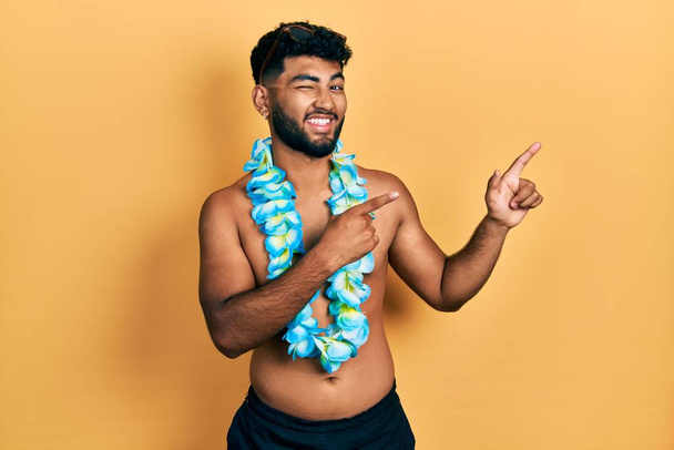 Homem árabe com barba vestindo maiô e lei havaiana sorrindo e olhando para a câmera apontando com duas mãos e dedos para o lado.  - Foto, Imagem