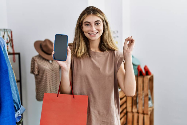 Young blonde woman holding shopping bags showing smartphone screen smiling happy pointing with hand and finger to the side  - Fotografie, Obrázek