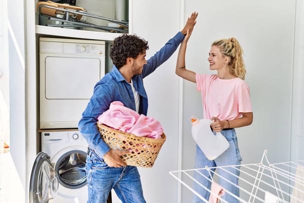 Young couple smiling happy holding laundry basket and detergent bottle high five at home. - Foto, Imagen