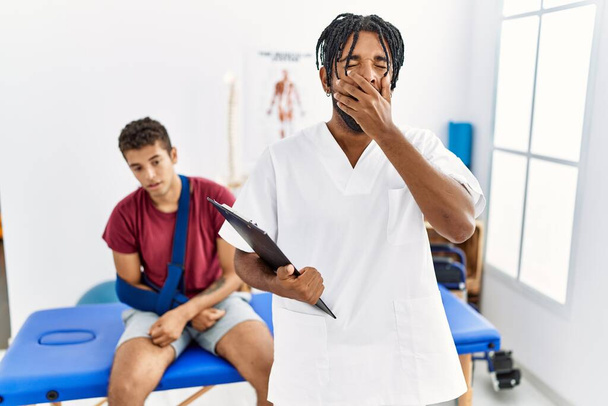 Young hispanic man working at pain recovery clinic with a man with broken arm bored yawning tired covering mouth with hand. restless and sleepiness.  - Photo, Image