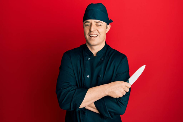 Beau jeune homme portant uniforme de cuisinier professionnel tenant couteau clin d'oeil en regardant la caméra avec une expression sexy, gai et heureux visage.  - Photo, image