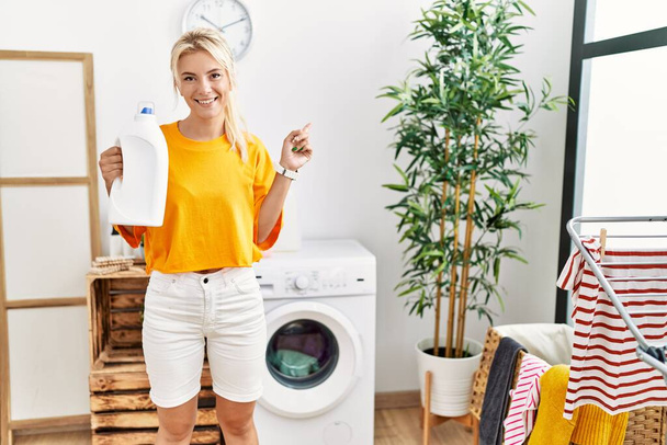 Nuori valkoihoinen nainen pyykkää pitäen pesuainetta pullon hymyillen onnellinen osoittaa käsi ja sormi sivulle  - Valokuva, kuva