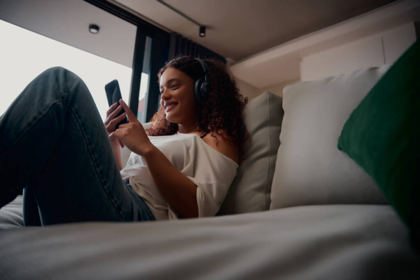 Mixed race female relaxing on bed texting boyfriend and listening to music smiling - Zdjęcie, obraz