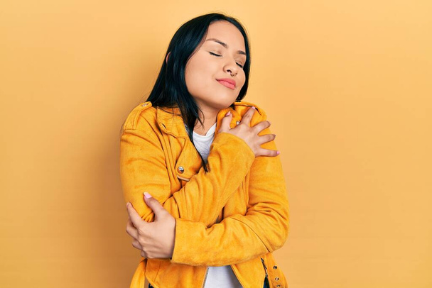 Beautiful hispanic woman with nose piercing wearing yellow leather jacket hugging oneself happy and positive, smiling confident. self love and self care  - Photo, Image