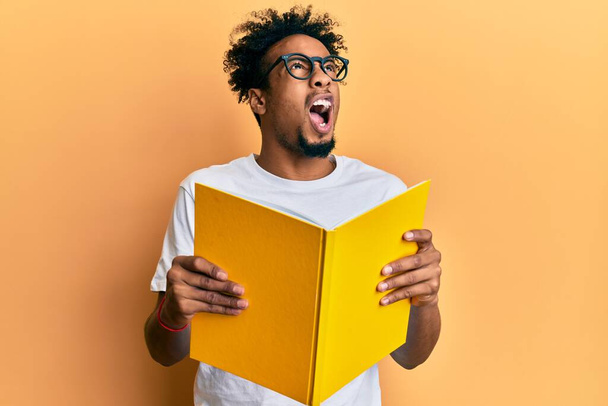 Joven afroamericano con barba leyendo un libro con gafas enojadas y locas gritando frustradas y furiosas, gritando de rabia mirando hacia arriba.  - Foto, imagen