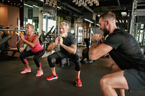 Active elderly woman and man performing squats in gym - Foto, imagen