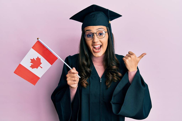 jonge latino vrouw draagt diploma-uitreiking uniform houden canada vlag wijzen duim naar de zijkant glimlachen blij met open mond  - Foto, afbeelding
