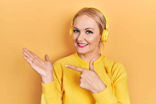 Nuori blondi nainen kuuntelee musiikkia kuulokkeilla hämmästynyt ja hymyilee kameralle samalla kun esittää käsi ja osoittaa sormella.  - Valokuva, kuva