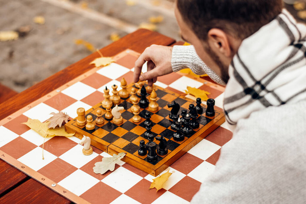 Üst manzara: Bir adam elinde piyon tutuyor ve oyun alanındaki bir sonbahar parkında bir masada satranç oynuyor. - Fotoğraf, Görsel