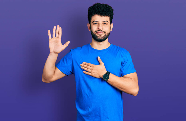 Giovane arabo con la barba indossa casual t shirt blu giurando con mano sul petto e palmo aperto, facendo un giuramento di fedeltà  - Foto, immagini