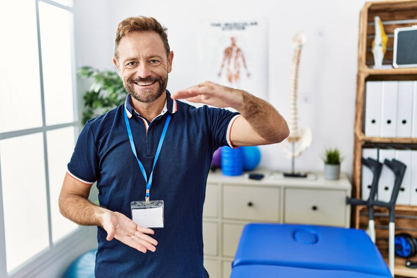 Hombre fisioterapeuta de mediana edad que trabaja en la clínica de recuperación del dolor gesticulando con las manos mostrando un signo de tamaño grande y grande, símbolo de medida. sonriendo mirando a la cámara. concepto de medición.  - Foto, Imagen