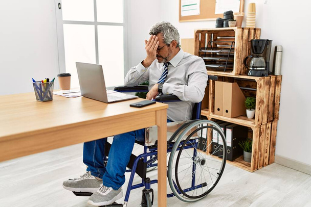 Middelbare leeftijd Spaanse man werkt op kantoor zitten op een rolstoel met droevige uitdrukking bedekken gezicht met handen tijdens het huilen. depressie concept.  - Foto, afbeelding