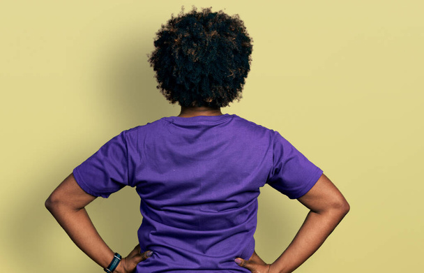 Femme afro-américaine aux cheveux afro portant un t-shirt décontracté violet debout vers l'arrière regardant loin avec les bras sur le corps  - Photo, image