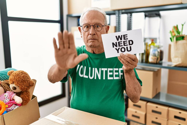 Hombre voluntario mayor sosteniendo necesitamos su pancarta con la mano abierta haciendo señal de stop con expresión seria y segura, gesto de defensa  - Foto, Imagen