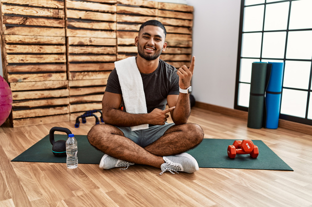 Hombre indio joven sentado en la colchoneta de entrenamiento en el gimnasio con una gran sonrisa en la cara, apuntando con el dedo de la mano hacia el lado mirando a la cámara.  - Foto, Imagen