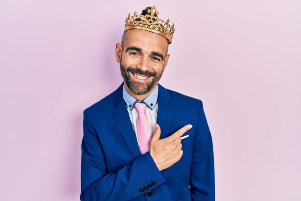 Jovem hispânico vestindo roupas de negócios e coroa rei sorrindo alegre apontando com a mão e o dedo para o lado  - Foto, Imagem