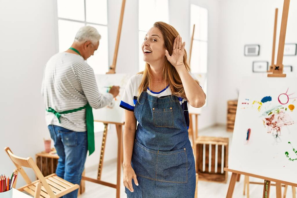 Hispanic woman wearing apron at art studio smiling with hand over ear listening an hearing to rumor or gossip. deafness concept.  - Foto, immagini