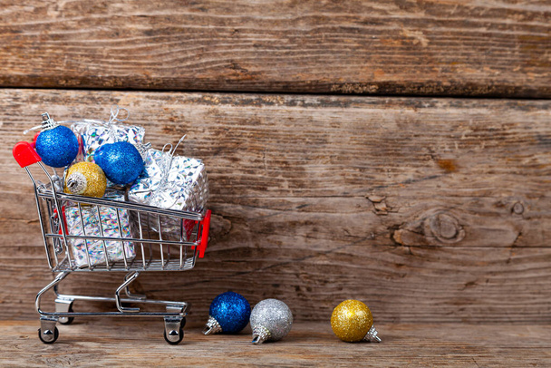 Shopping cart with Christmas gifts and balls on a wooden background. Holiday sale. - Fotografie, Obrázek