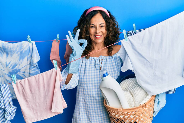 中世のヒスパニック女性を保持洗濯バスケット洗濯服で服を着て指でokサインをします,笑顔優しいジェスチャー優れたシンボル  - 写真・画像