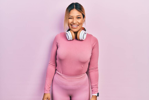 Beautiful hispanic woman wearing gym clothes and using headphones winking looking at the camera with sexy expression, cheerful and happy face.  - Photo, Image