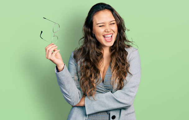 Young hispanic girl wearing business clothes holding glasses winking looking at the camera with sexy expression, cheerful and happy face.  - Photo, Image