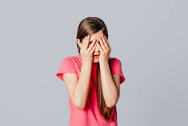 Испуганная, встревоженная молодая девушка изумляла потрясенные руки закрывающими лицо, носила случайную розовую футболку, стоя на сером фоне. Молодая эмоциональная женщина - Фото, изображение