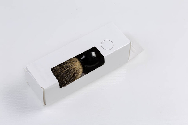 Новая щетка для бритья с натуральной щетиной барсука и пластиковой ручкой в картонной упаковке лежит на светлой поверхности - Фото, изображение