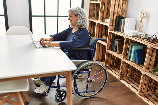Μέση ηλικία γκρίζα μαλλιά με αναπηρία γυναίκα χρησιμοποιώντας φορητό υπολογιστή κάθεται σε αναπηρική καρέκλα στο σπίτι. - Φωτογραφία, εικόνα