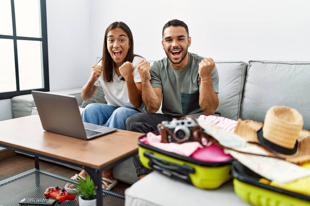 Jeune couple interracial emballant des vêtements d'été dans une valise en regardant un ordinateur portable crier fier, célébrer la victoire et le succès très excité avec les bras levés  - Photo, image