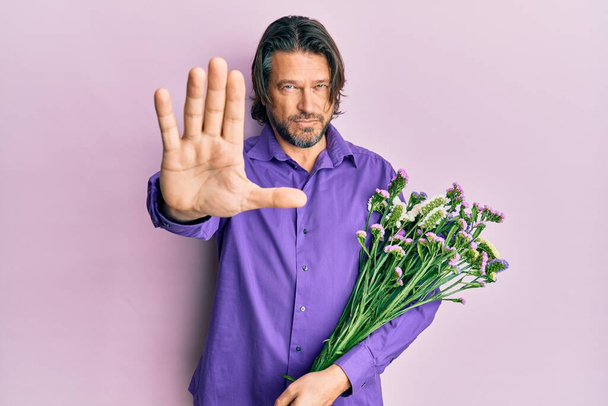 Moyen-âge bel homme tenant des fleurs avec la main ouverte faisant stop signe avec expression sérieuse et confiante, geste de défense  - Photo, image
