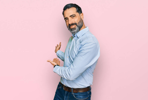 Mann mittleren Alters mit Bart und Business-Hemd lädt ein, lächelnd und natürlich mit offener Hand einzutreten  - Foto, Bild