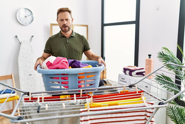 Middle age man doing laundry holding basket in shock face, looking skeptical and sarcastic, surprised with open mouth  - Foto, imagen