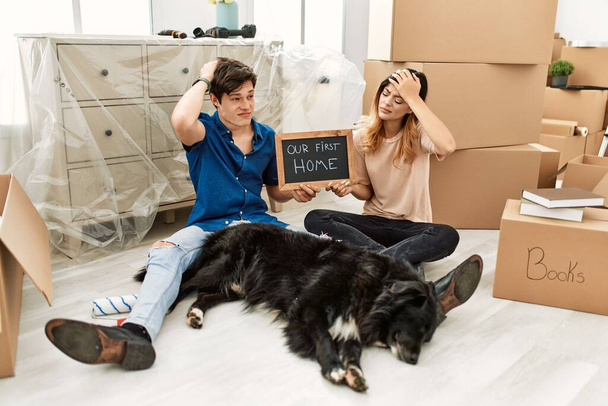 Young caucasian couple with dog holding our first home blackboard at new house confuse and wondering about question. uncertain with doubt, thinking with hand on head. pensive concept.  - Фото, изображение