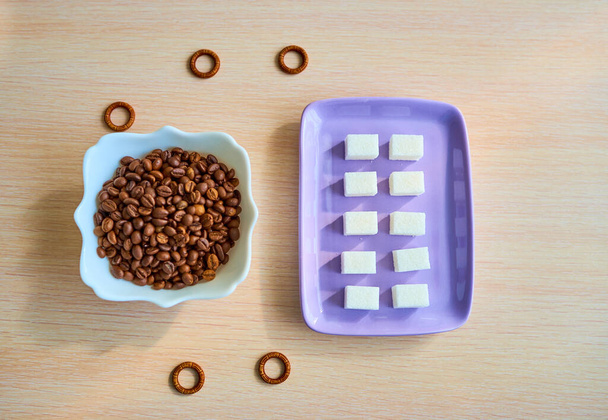 На столі лежить фіолетова тарілка з десятьма шматочками білого цукру та білою тарілкою з купою кавових зерен. Вид зверху
 - Фото, зображення