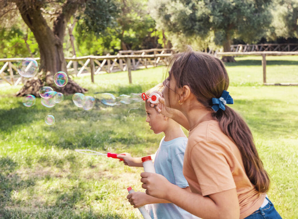 Hermano y hermana soplando burbujas de jabón al aire libre. Linda niña y niño divirtiéndose en un parque en verano. Concepto de actividad al aire libre para niños, auténtico momento de infancia. Camiseta maqueta - Foto, imagen