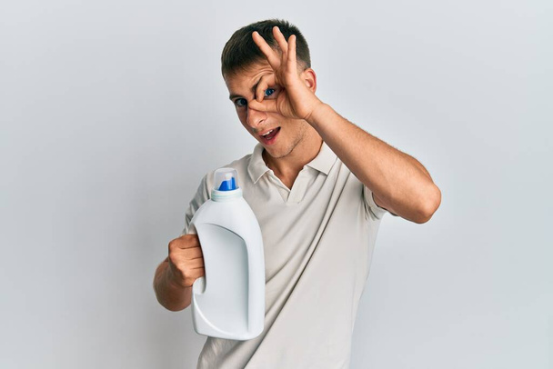Молодий білий чоловік тримає пляшку миючого засобу усміхненим щасливим роблячи знак ОК з рукою на око дивиться крізь пальці
  - Фото, зображення