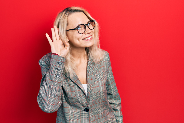 Красивая кавказская блондинка в деловой куртке и в очках, улыбаясь рукой за ухом, слушая и слушая слухи или сплетни. концепция глухоты.  - Фото, изображение