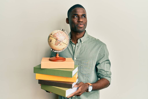 Jonge Afro-Amerikaanse man met vintage wereldbal en boeken glimlachend naar de zijkant kijkend en wegdenkend.  - Foto, afbeelding