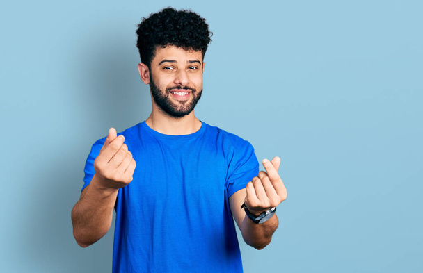 Jeune homme arabe avec barbe vêtu d'un t-shirt bleu décontracté faisant un geste d'argent avec les mains, demandant un paiement de salaire, entreprise millionnaire  - Photo, image