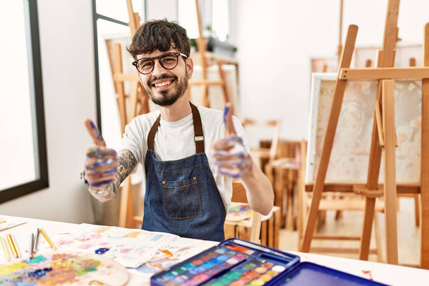 Homme hispanique avec barbe au studio d'art approuvant faire un geste positif avec la main, pouces levés souriant et heureux pour le succès. geste gagnant.  - Photo, image