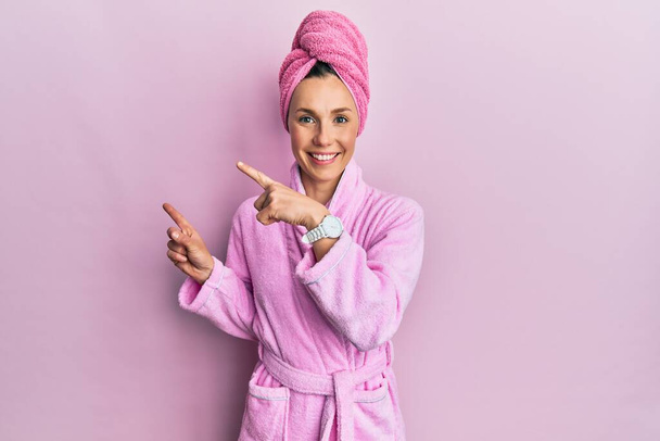 Jeune femme blonde portant un bonnet de serviette de douche et un peignoir souriant et regardant la caméra pointant avec deux mains et doigts sur le côté.  - Photo, image