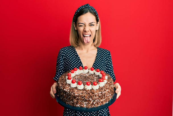 Joven mujer rubia caucásica sosteniendo pastel de chocolate sacando la lengua feliz con expresión divertida.  - Foto, imagen