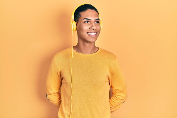 Jeune homme afro-américain écoutant de la musique à l'aide d'écouteurs détournant les yeux avec le sourire sur le visage, expression naturelle. riant confiant.  - Photo, image