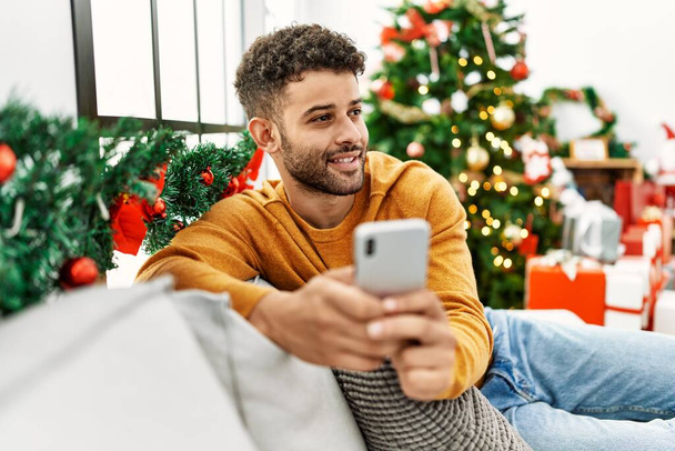 Hombre árabe joven usando teléfono inteligente sentado en el sofá por árbol de Navidad en casa. - Foto, Imagen
